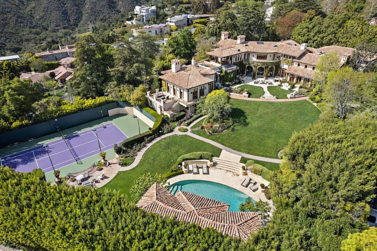 This expansive aerial view captures a luxurious Mediterranean-style estate nestled in a lush, hilly landscape. The property features a sprawling main residence with a terracotta-tiled roof, a large manicured lawn, a private tennis court, and a swimming pool with an adjacent pool house. The perspective provides a comprehensive look at the property's generous outdoor living spaces and its integration into the surrounding verdant hillside environment.