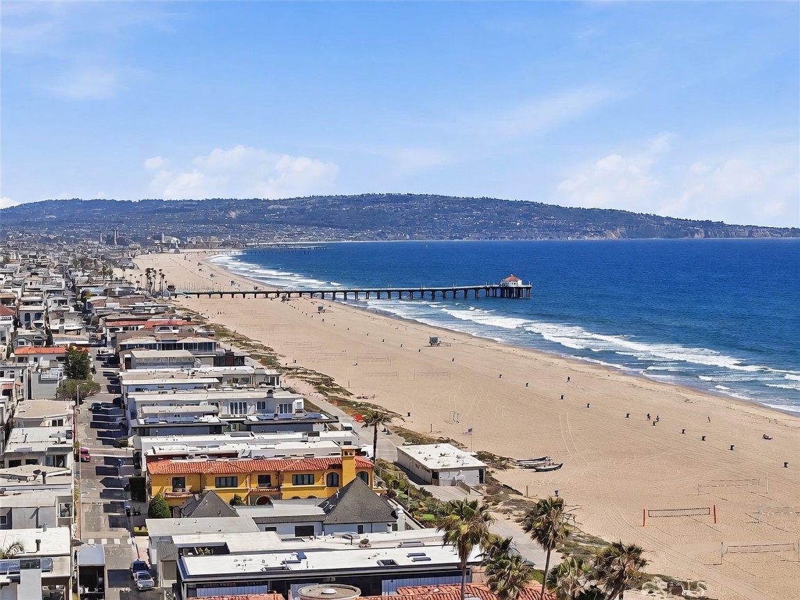 This high-angle aerial view captures a stunning coastal scene featuring a wide sandy beach stretching along the Pacific Ocean. A long pier extends into the blue water, while the foreground showcases a dense neighborhood of residential homes with varied architectural styles. The perspective provides a clear sense of the property's prime location near the shoreline and the expansive horizon.