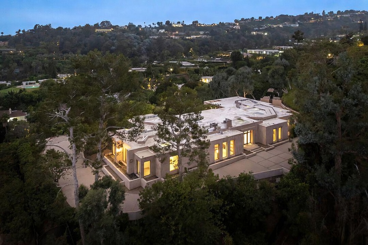 This high-angle aerial shot captures a sprawling, modern luxury estate nestled within a lush, hilly landscape at twilight. The architecture features clean, geometric lines and a flat roof, with warm interior lighting glowing through large windows, creating an inviting contrast against the cool blue tones of the evening sky. The property is surrounded by dense greenery and mature trees, emphasizing a sense of privacy and exclusivity in a prestigious residential setting.