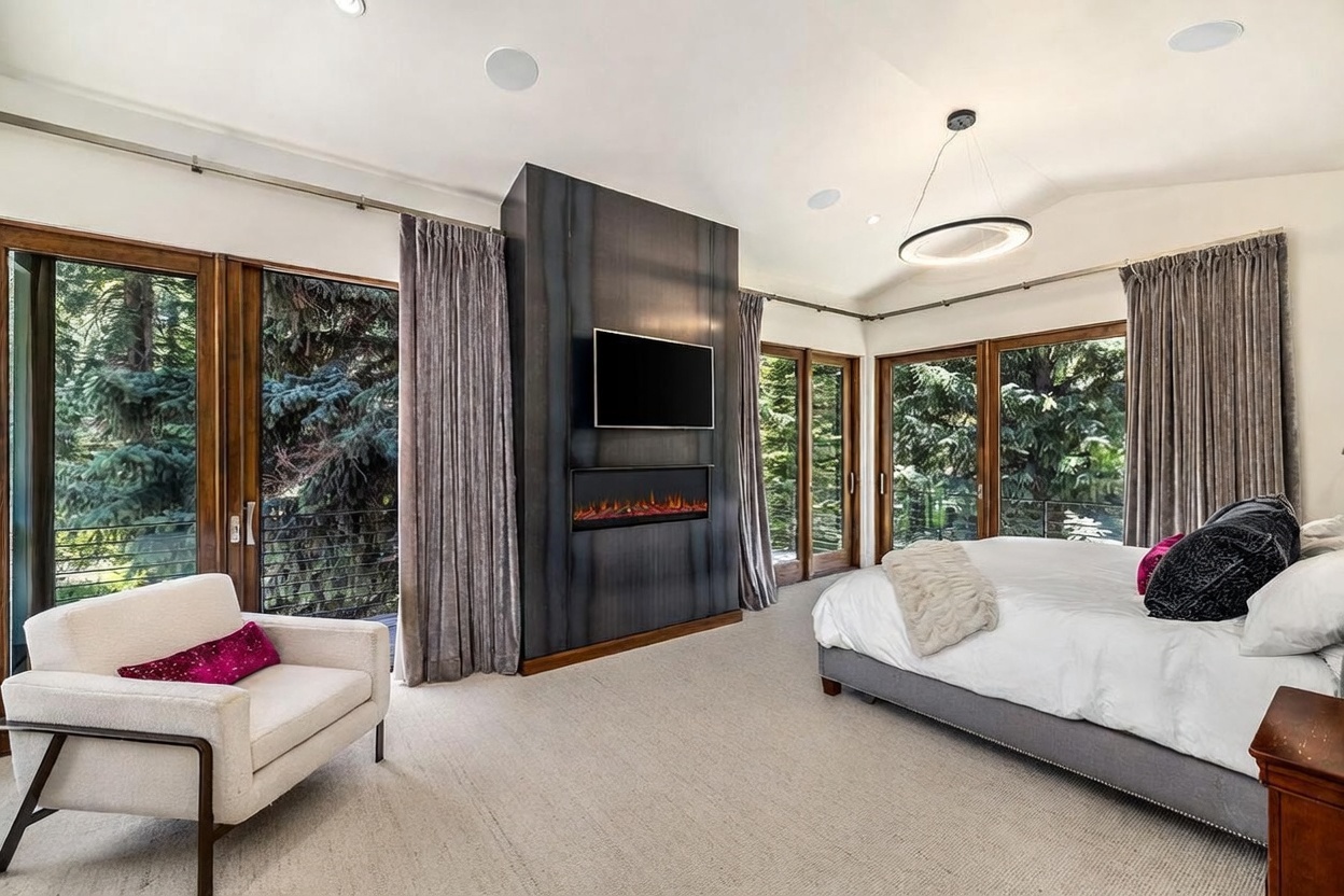 This spacious primary bedroom features a modern aesthetic with a prominent dark, metallic-finished chimney breast housing a television and a linear fireplace. The room is anchored by a large bed with plush bedding and is flanked by expansive windows that offer serene views of the surrounding forest. A comfortable armchair sits in the corner, and the neutral color palette creates a calm, sophisticated atmosphere.