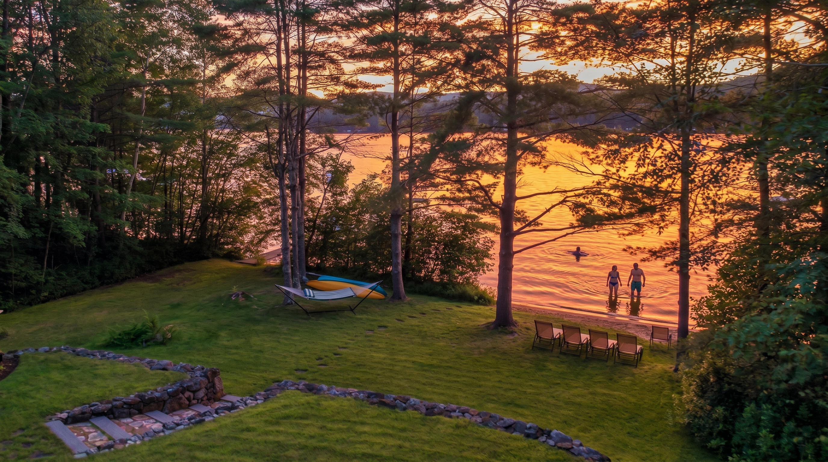 This elevated perspective captures a serene, grassy backyard sloping gently toward a tranquil lake at sunset. The scene features a hammock nestled between mature trees, a set of lounge chairs positioned near the water's edge, and a stone-retaining wall with integrated steps leading down the lawn. The warm, golden light of the setting sun reflects off the water, creating a peaceful and inviting atmosphere for outdoor relaxation.