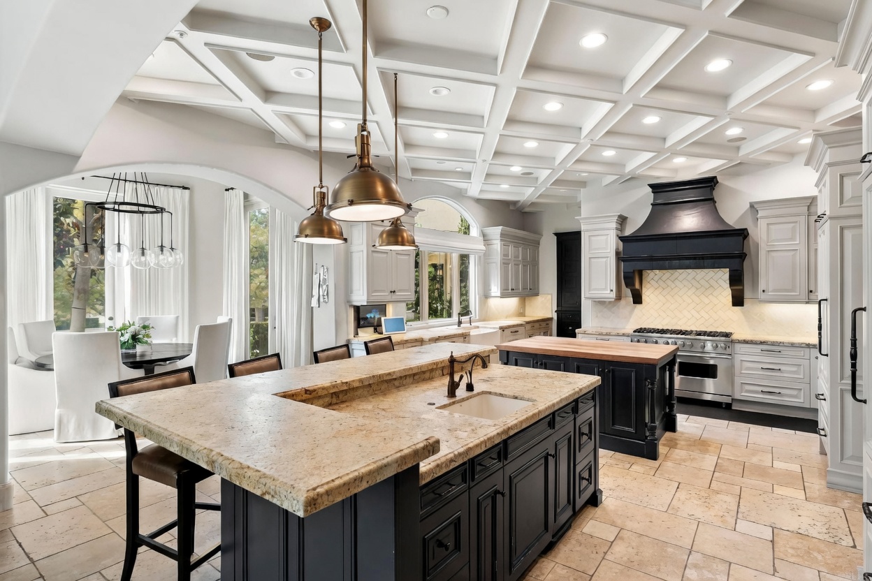 This is a well-lit, luxurious kitchen featuring a large island with a beige countertop and dark cabinetry, complemented by pendant lighting. The kitchen also includes a secondary island, stainless steel appliances, and a black range hood, all set against a backdrop of white cabinetry and beige tile flooring. The perspective is a wide shot, showcasing the spaciousness and high-end finishes of the kitchen.