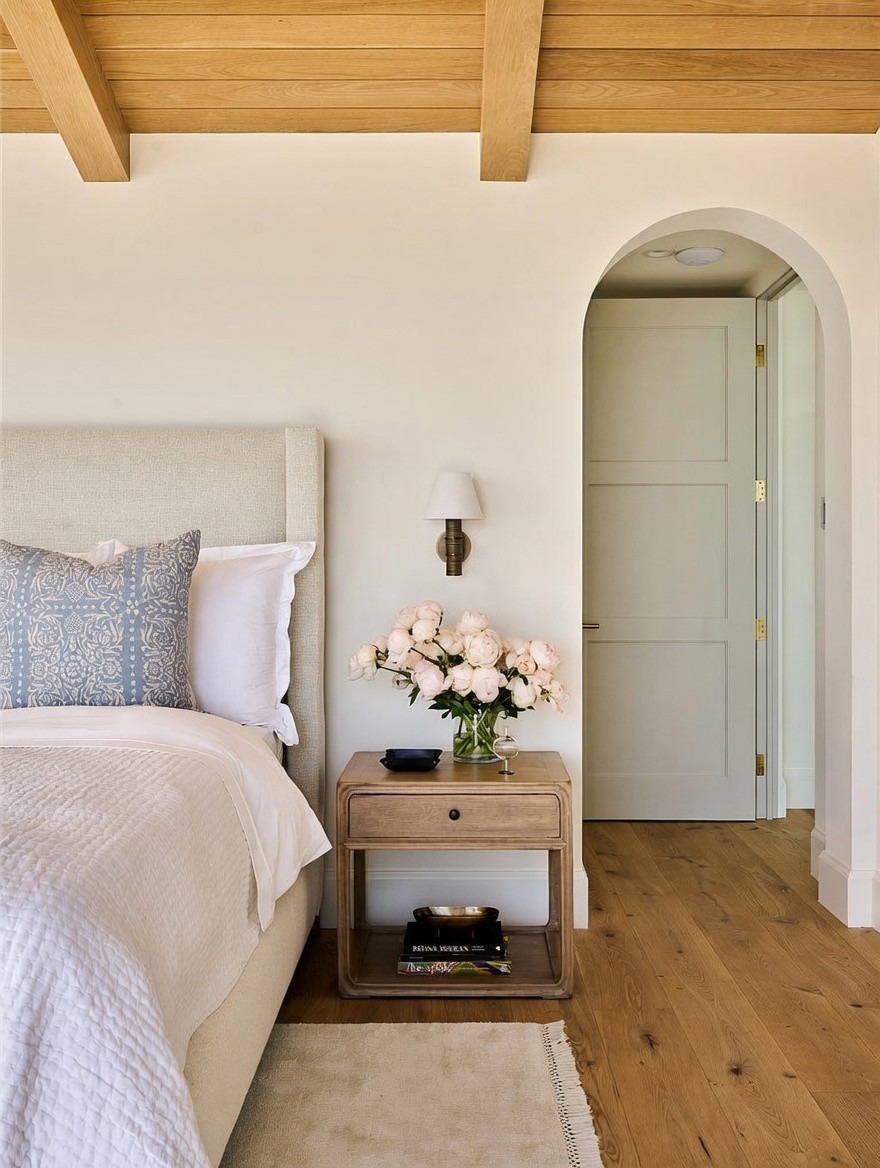 This serene bedroom features a neutral color palette with a textured upholstered headboard and light wood flooring. A bedside table holds a vase of fresh peonies and a wall-mounted sconce, while an arched doorway leads to an adjacent room. The space is accented by a wood-paneled ceiling, creating a warm and inviting atmosphere.