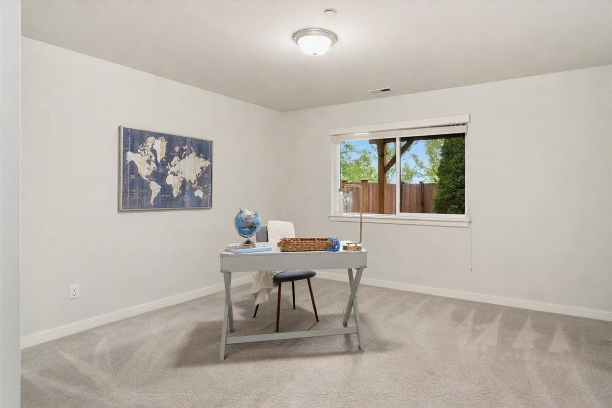 This bright and minimalist home office features neutral walls, plush carpeting, and a central desk setup facing a large window that provides natural light and a view of the outdoors. A world map wall art piece adds a decorative touch, while the clean, uncluttered layout creates a productive and serene atmosphere. The perspective is centered, highlighting the functional simplicity of the workspace.