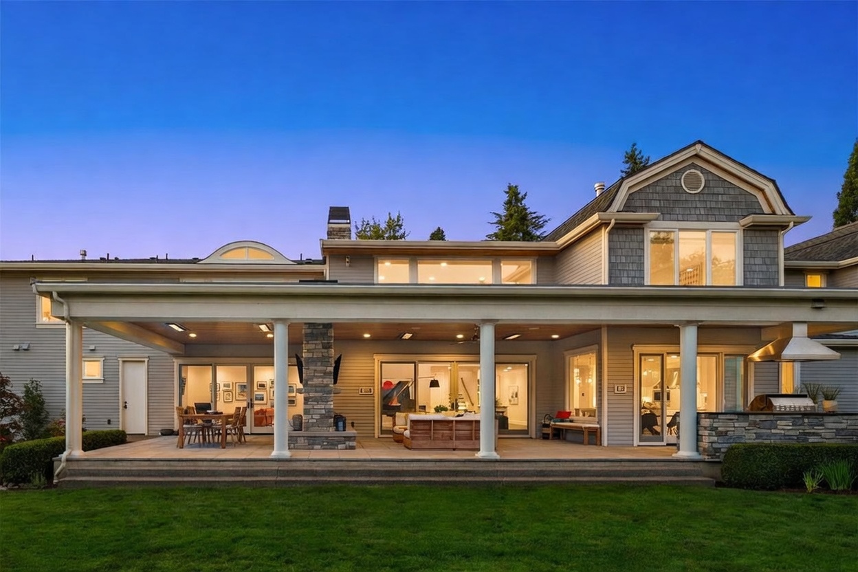 This rear view of a large, multi-story home showcases a spacious covered patio area perfect for outdoor entertaining. The patio features a stone-clad chimney breast, recessed lighting, and ample space for dining and lounge furniture, all overlooking a well-manicured lawn. The exterior is finished in horizontal siding with a mix of shingle-style siding on the upper gable, creating a warm and inviting residential aesthetic.