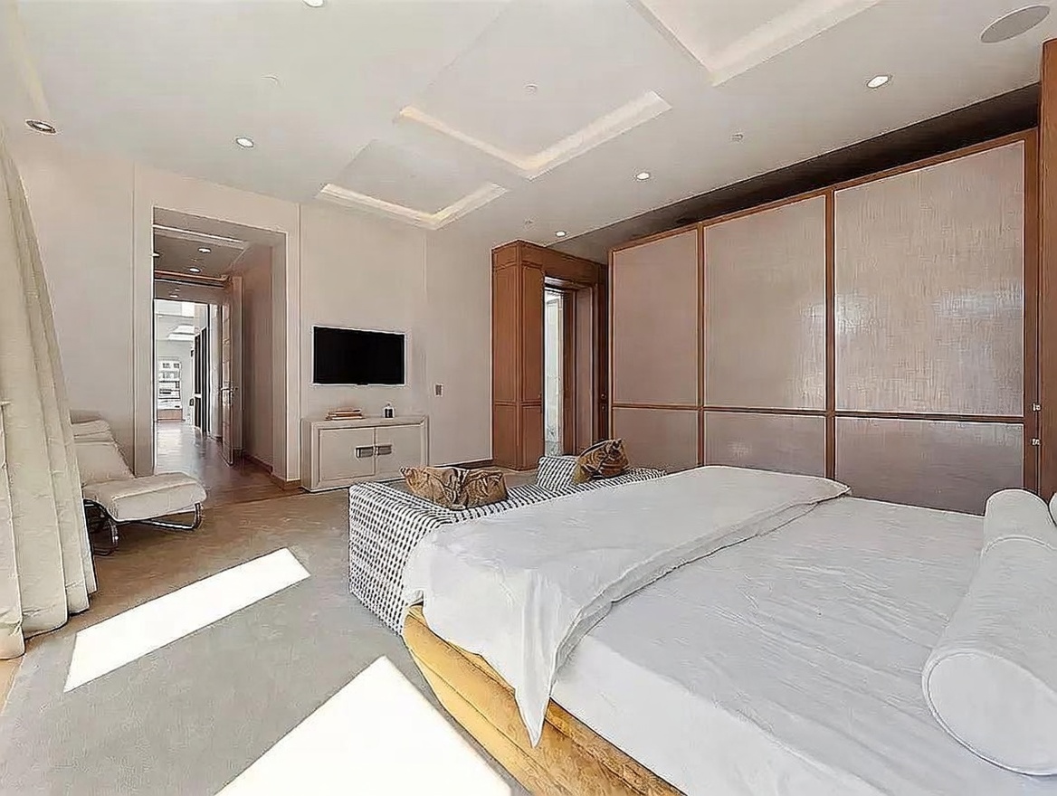 This spacious primary bedroom features a large bed with a patterned bench at the foot and a modern chaise lounge positioned near a tall window. The room is accented by a large, multi-panel wardrobe with textured glass doors and a recessed ceiling design with integrated lighting. A television is mounted above a sleek, light-colored media console, and the neutral color palette creates a serene and sophisticated atmosphere.