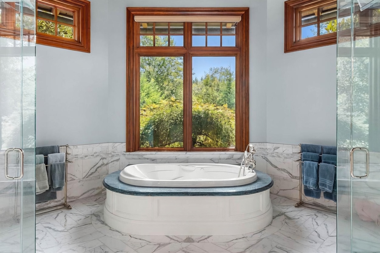 This primary bathroom features a central soaking tub set against a large window with a view of the surrounding trees. The space is finished with light-toned walls, patterned stone flooring, and a stone-topped tub surround, flanked by glass-enclosed shower areas on either side. The room is accented by warm wood-framed windows and metal towel racks, creating a bright and serene atmosphere.