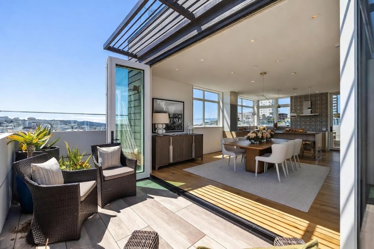 This image captures a seamless transition between an indoor dining area and an expansive outdoor balcony, highlighting an open-concept living space. The balcony features wicker-style seating and potted plants, while the interior showcases a wooden dining table, modern chairs, and a kitchen area with patterned wall tiles. Large sliding glass doors create an airy, sophisticated atmosphere with panoramic city views in the background.