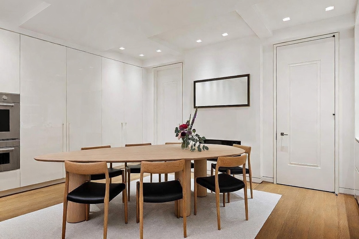 This bright and minimalist dining room features a large, oval-shaped light wood table surrounded by six modern chairs with black upholstered seats. The space is defined by clean white walls, recessed ceiling lighting, and a light-colored area rug placed over hardwood floors. A rectangular mirror hangs on the wall above a low-profile console, creating a sophisticated and airy atmosphere.