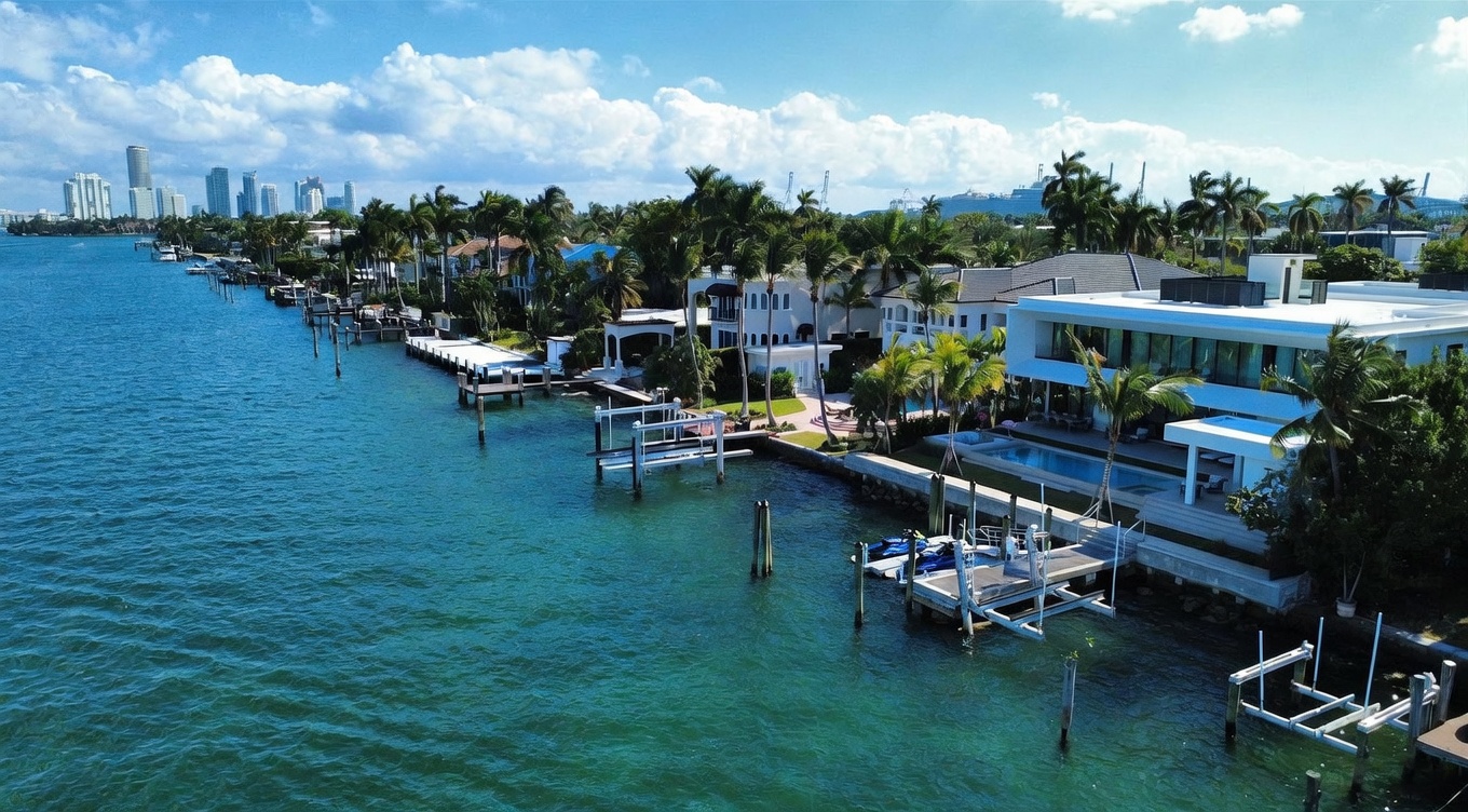 This high-angle aerial view captures a luxurious waterfront estate featuring a modern white residence with a private pool and multiple boat docks extending into the turquoise bay. The property is surrounded by lush tropical landscaping, including numerous palm trees, and offers a clear view of the distant city skyline across the water. The scene conveys an upscale, serene coastal lifestyle with direct access to the waterway.