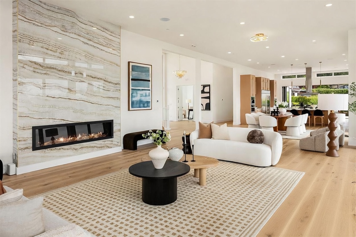 This spacious, open-concept living area features a prominent stone-clad chimney breast with a linear fireplace as its focal point. The room is furnished with a curved white sofa, matching armchairs, and a layered coffee table arrangement atop a textured area rug, creating a sophisticated and inviting atmosphere. In the background, the space flows seamlessly into a dining area and kitchen, characterized by light wood flooring and a bright, airy aesthetic.