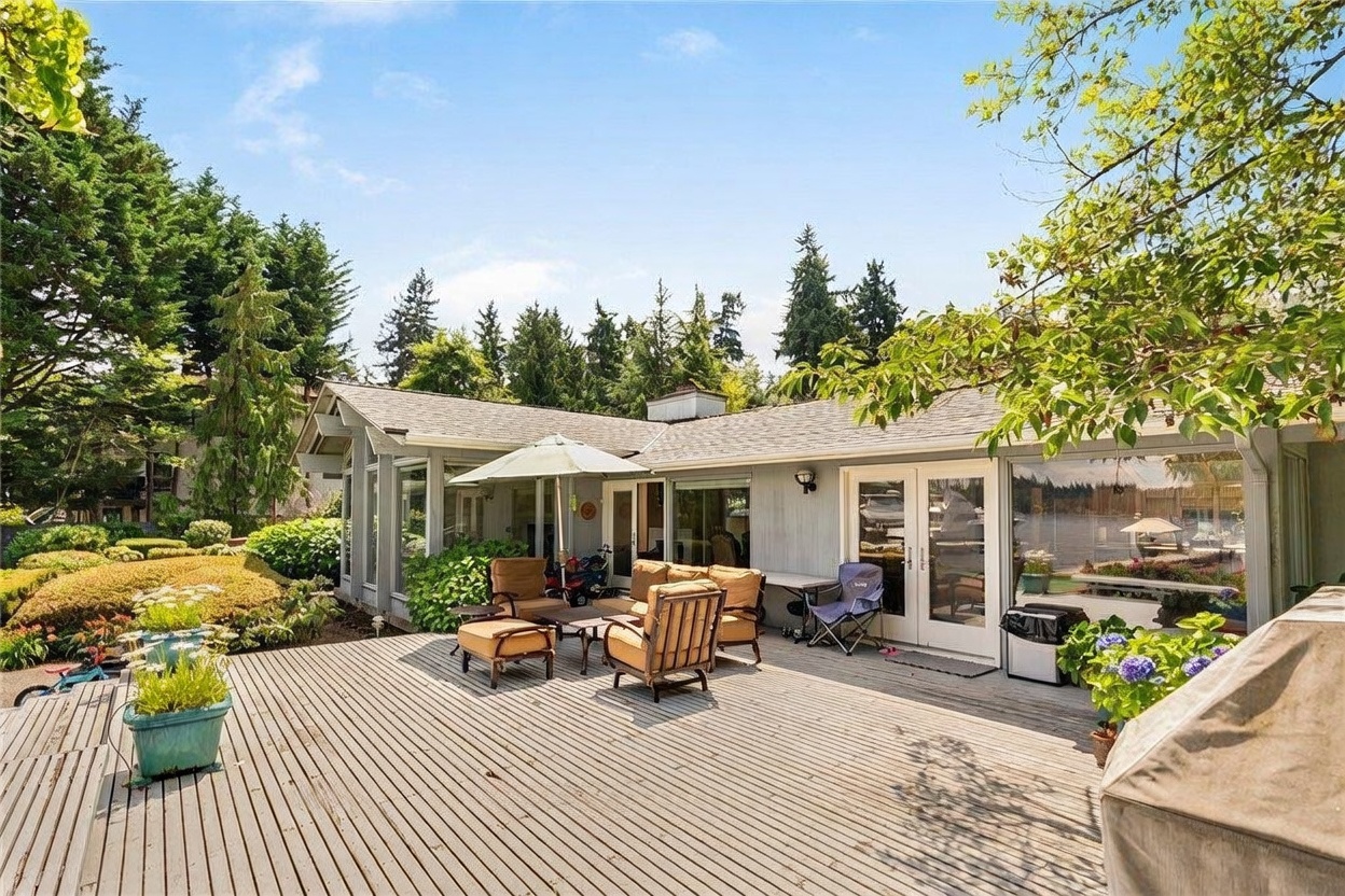 This image captures a spacious wooden deck attached to the rear of a single-story home, featuring a comfortable outdoor seating area with a patio umbrella. The deck overlooks a serene waterfront setting, framed by lush green trees and a well-maintained garden. The perspective is from the deck looking toward the house, highlighting the seamless transition between indoor and outdoor living spaces.