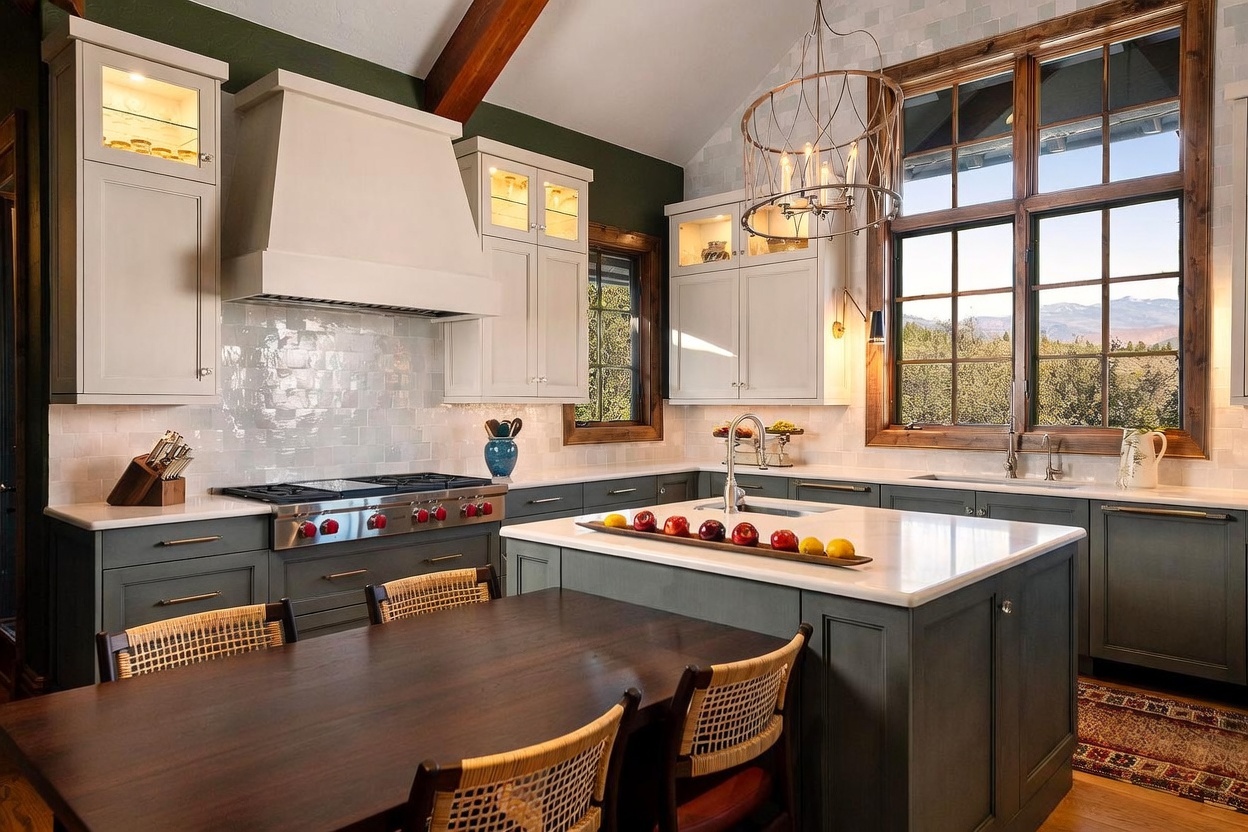 This elegant kitchen features a two-tone color palette with dark lower cabinetry and light upper cabinets, complemented by a large central island topped with a light-colored stone surface. A professional-grade range with a custom hood serves as the focal point, while a large window above the sink provides ample natural light and scenic outdoor views. The space is finished with a wooden dining table in the foreground and a decorative chandelier, creating a warm and inviting atmosphere.