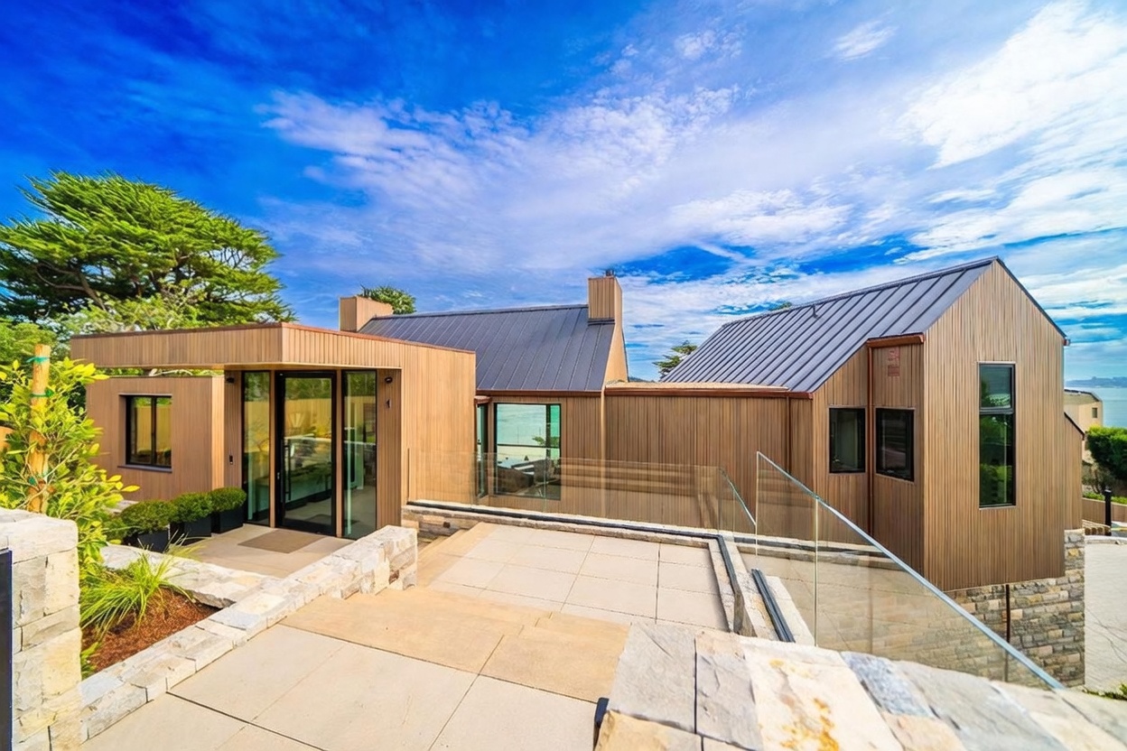 This modern architectural home features a striking entryway characterized by clean lines, vertical wood-toned siding, and a dark metal roof. A tiered stone patio leads to a glass-paneled front door, complemented by a sleek glass railing that overlooks the surrounding landscape. The composition captures a bright, sunny day with a clear blue sky, emphasizing the home's contemporary design and seamless indoor-outdoor transition.