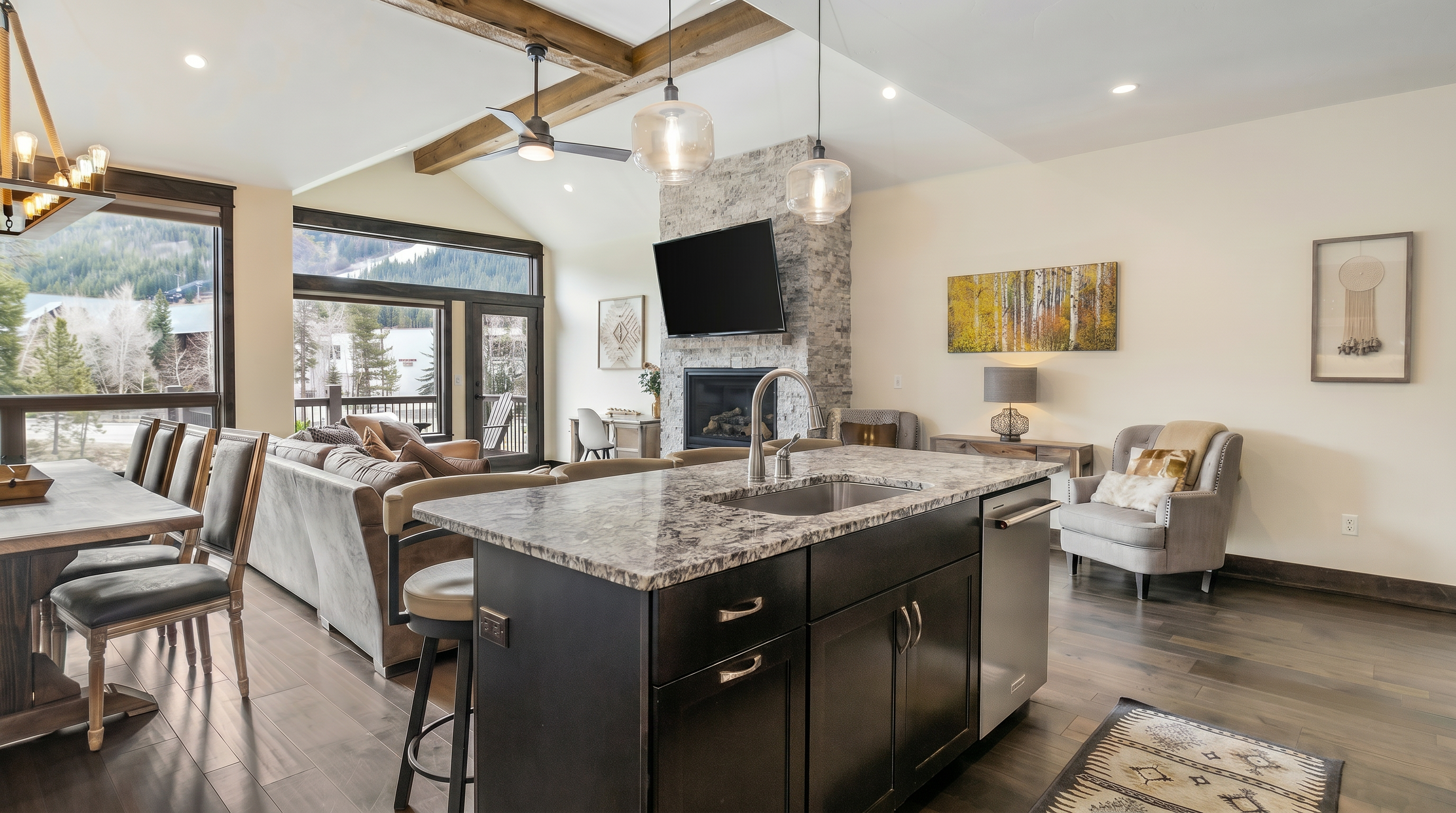 This open-concept kitchen features a large dark-wood island with a granite countertop, serving as the focal point of the space. The room flows seamlessly into a cozy living area with a stone fireplace, vaulted ceilings with exposed wooden beams, and expansive windows that offer scenic mountain views. The interior design blends rustic mountain charm with modern amenities, creating a warm and inviting atmosphere.
