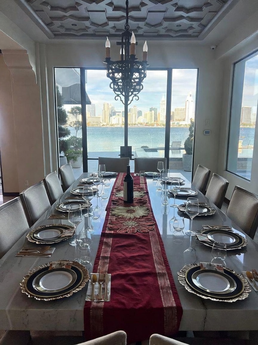 This elegant dining room features a long, light-toned stone table set for ten, centered under a large, ornate black metal chandelier. The room is framed by expansive windows that offer a stunning, unobstructed view of a bay and a distant city skyline. A deep red table runner adds a pop of color to the sophisticated, neutral-toned space, which is topped by a detailed, geometric coffered ceiling.