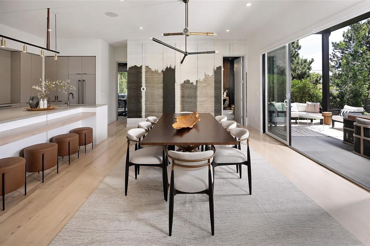 This open-concept dining area features a dark wood table surrounded by modern upholstered chairs, set atop a textured area rug. To the left, a kitchen island with leather-topped stools provides additional seating, while a large sliding glass door on the right opens to an outdoor patio. The space is defined by a striking abstract wall mural and a contemporary geometric chandelier, creating a sophisticated and airy atmosphere.