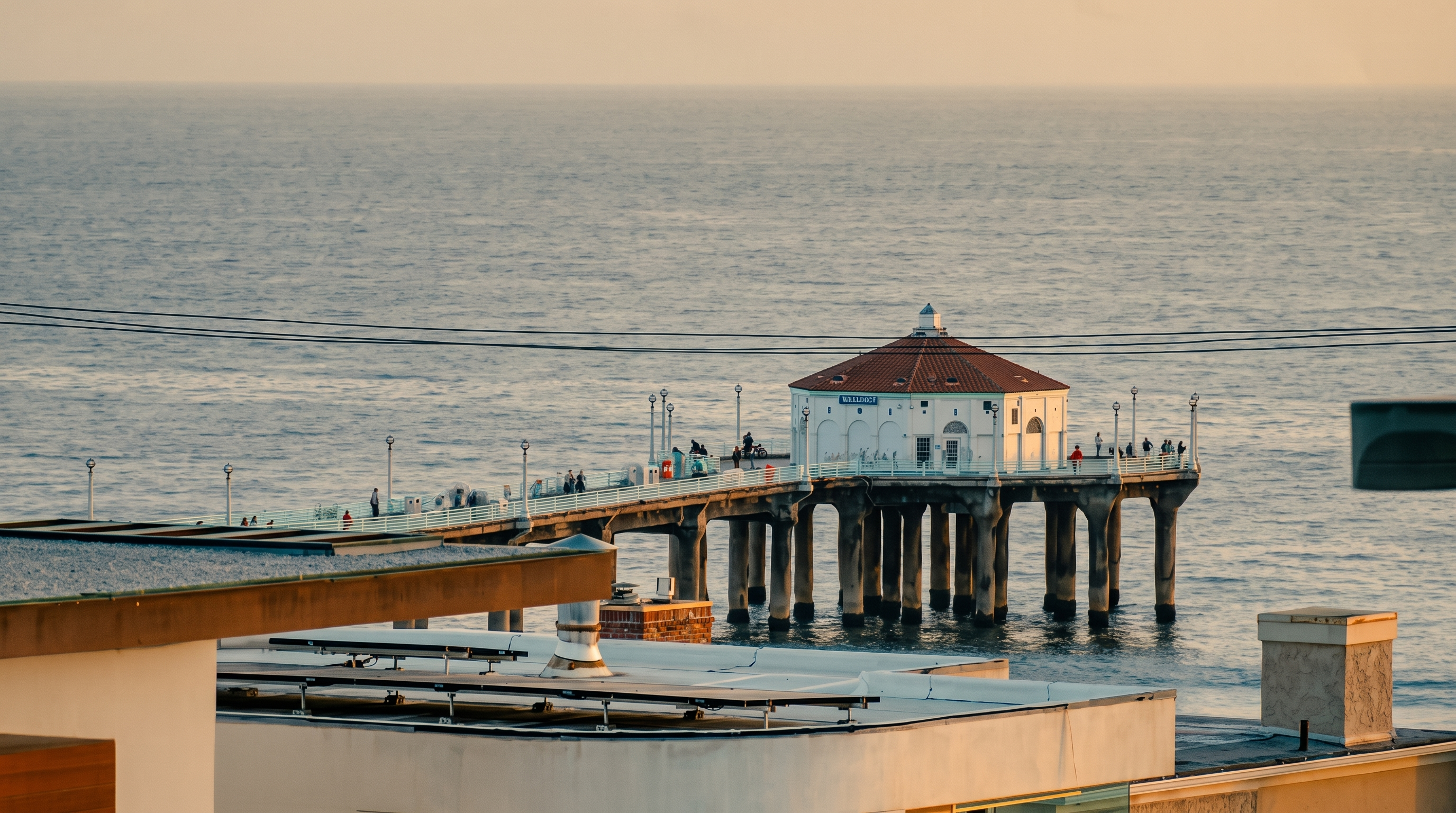 This elevated, wide-angle shot captures a scenic view of the Manhattan Beach Pier extending into the Pacific Ocean. In the foreground, the rooftops of nearby coastal buildings are visible, providing a sense of the property's proximity to the landmark. The scene is bathed in the soft, warm light of late afternoon, highlighting the pier's iconic round building and the calm, expansive horizon.