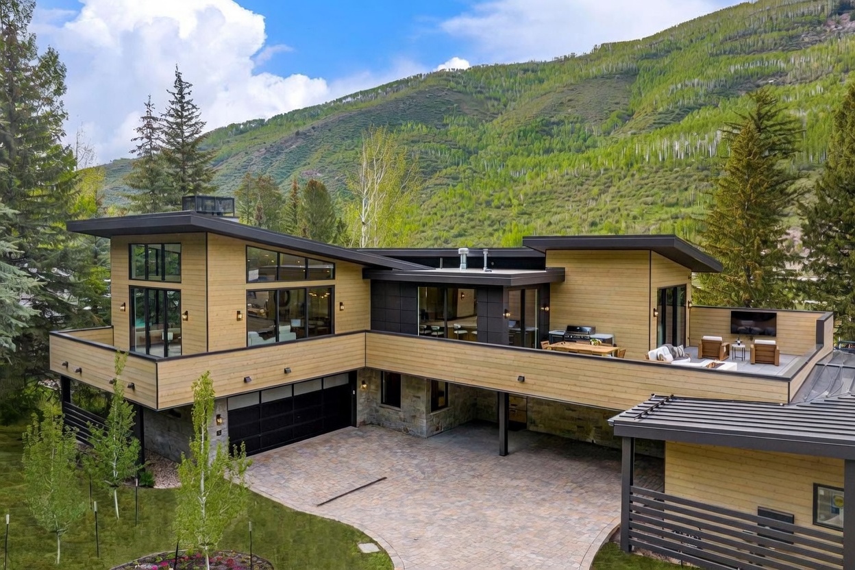 This elevated perspective captures a modern mountain residence featuring a striking mix of horizontal wood siding, dark metal accents, and stone masonry. The home boasts a spacious upper-level deck equipped with outdoor seating and a grill, overlooking a paved driveway and garage area. Set against a backdrop of lush, tree-covered hills, the architectural design emphasizes clean lines and a seamless integration with the natural landscape.