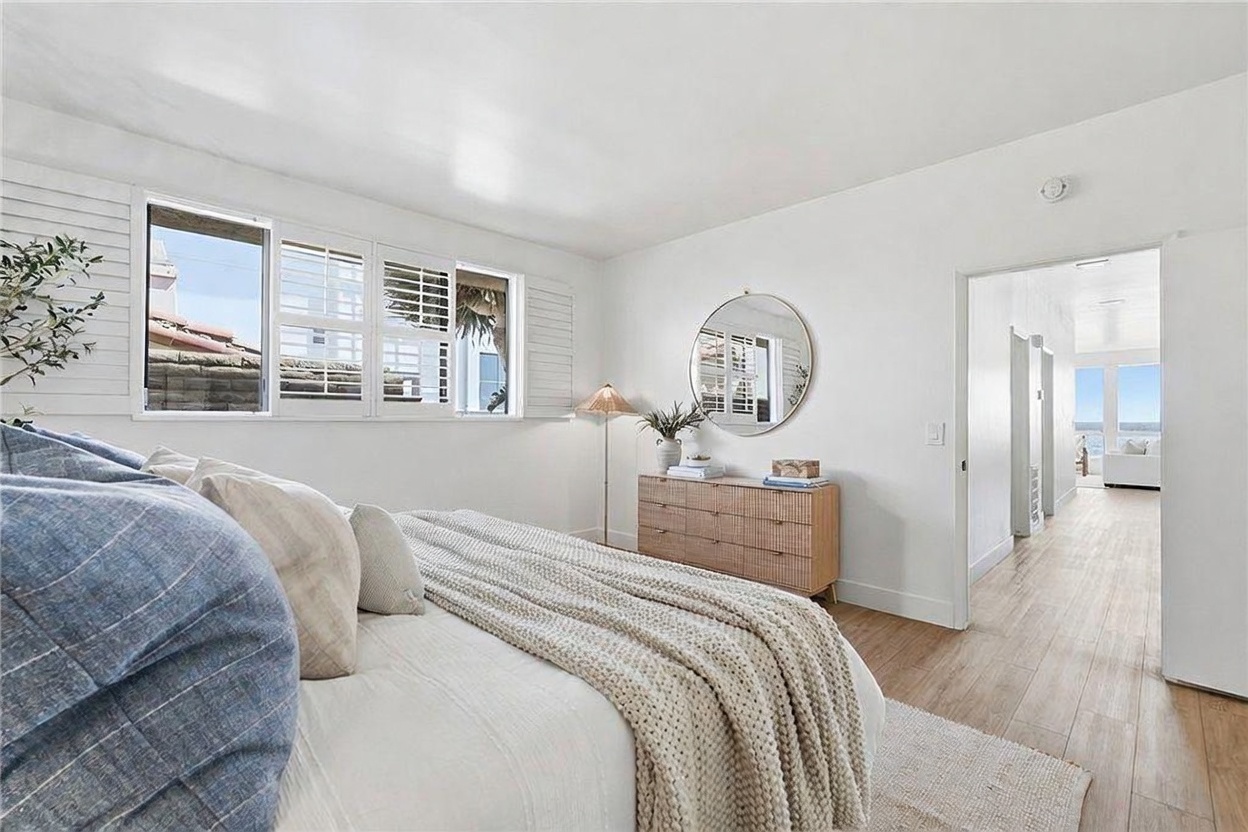 This bright and airy primary bedroom features light wood-toned flooring and crisp white walls, creating a serene coastal aesthetic. A plush bed with a textured throw blanket sits in the foreground, while a round mirror hangs above a wooden dresser, reflecting the room's clean lines. A doorway leads into a sun-drenched hallway that offers a glimpse of the ocean view beyond, emphasizing the home's open and connected layout.