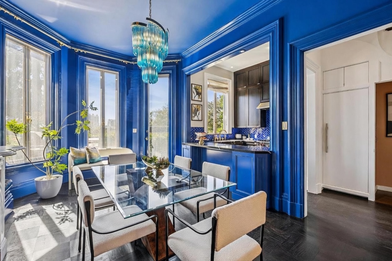 This vibrant dining room features bold, high-gloss blue walls and trim, creating a dramatic and sophisticated atmosphere. A glass-topped table is surrounded by modern, light-toned upholstered chairs, positioned beneath a striking, multi-tiered blue glass chandelier. The space flows seamlessly into an adjacent kitchen area, with dark wood flooring and large windows that flood the room with natural light.