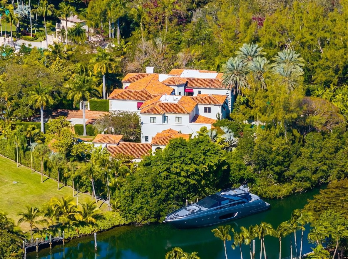 This high-angle aerial view captures a sprawling Mediterranean-style estate nestled within a lush, tropical landscape. The property features a multi-gabled roof with terracotta tiles, white stucco walls, and a private dock on a calm canal where a large, dark-hulled yacht is moored. The surrounding grounds are densely populated with mature palm trees and vibrant greenery, creating a secluded and luxurious waterfront atmosphere.
