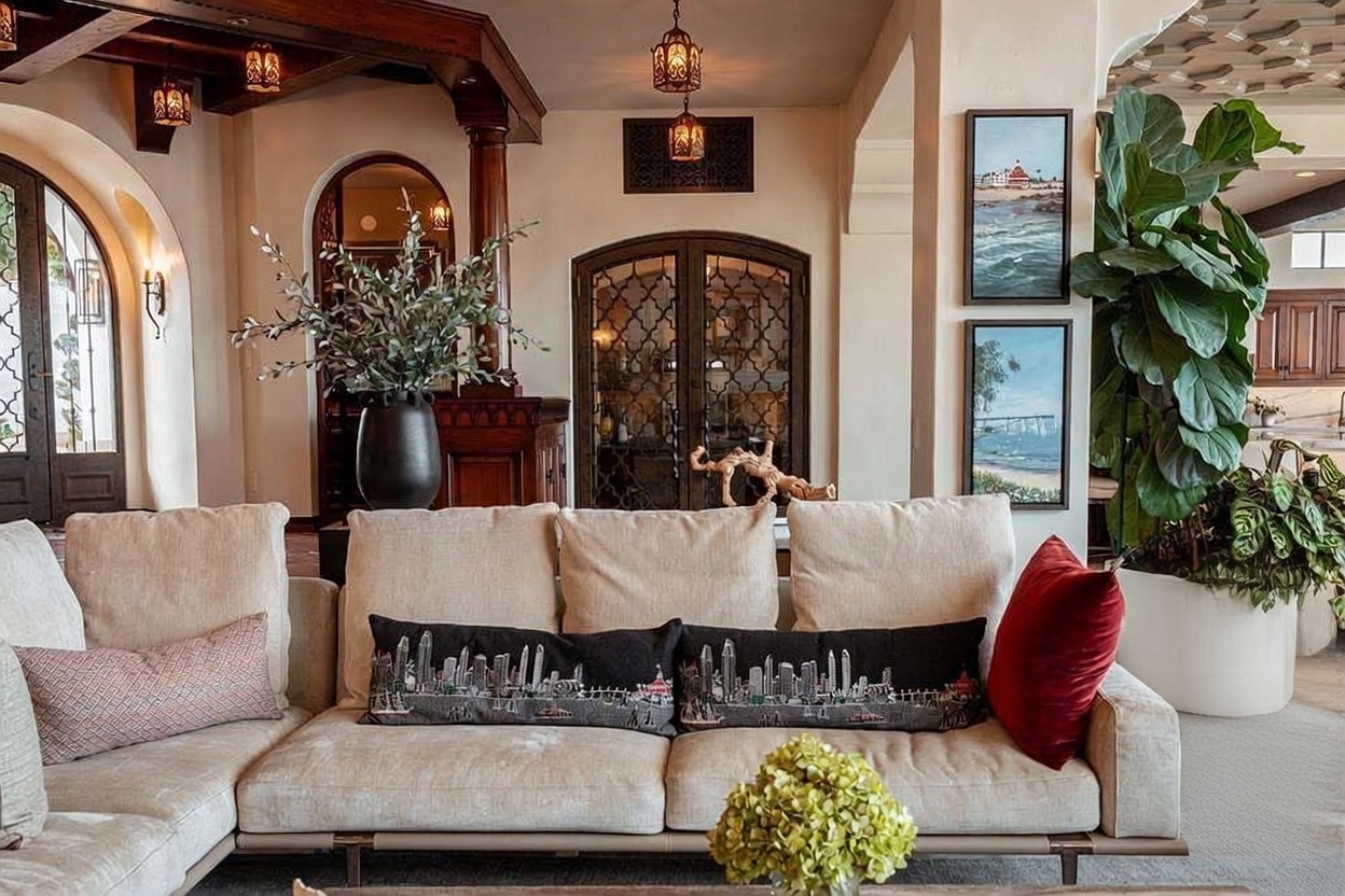 This elegant living room features a large, neutral-toned sectional sofa adorned with decorative pillows, including two featuring a detailed city skyline motif. The space is anchored by a dark, ornate wooden cabinet and a sophisticated arched doorway with intricate metal lattice work, creating a warm, Mediterranean-inspired atmosphere. Lush greenery, including a large potted fiddle leaf fig and a vase of olive branches, adds a vibrant organic touch to the refined interior design.