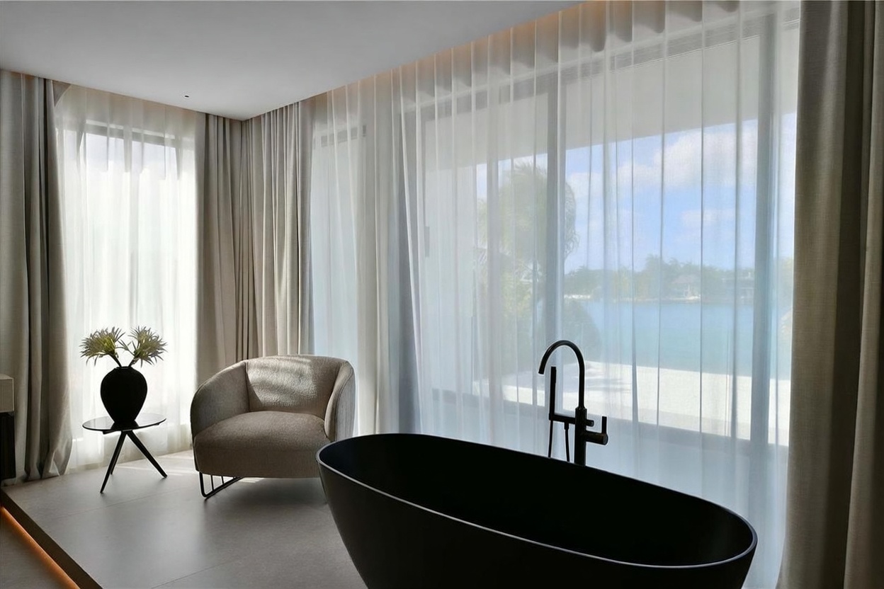 This sophisticated primary bathroom features a sleek, matte black freestanding soaking tub positioned as a focal point. A plush, curved armchair sits nearby on a minimalist platform, complemented by a small side table with a decorative plant. Large, floor-to-ceiling windows draped in sheer curtains provide soft, diffused light and a serene view of the water outside.