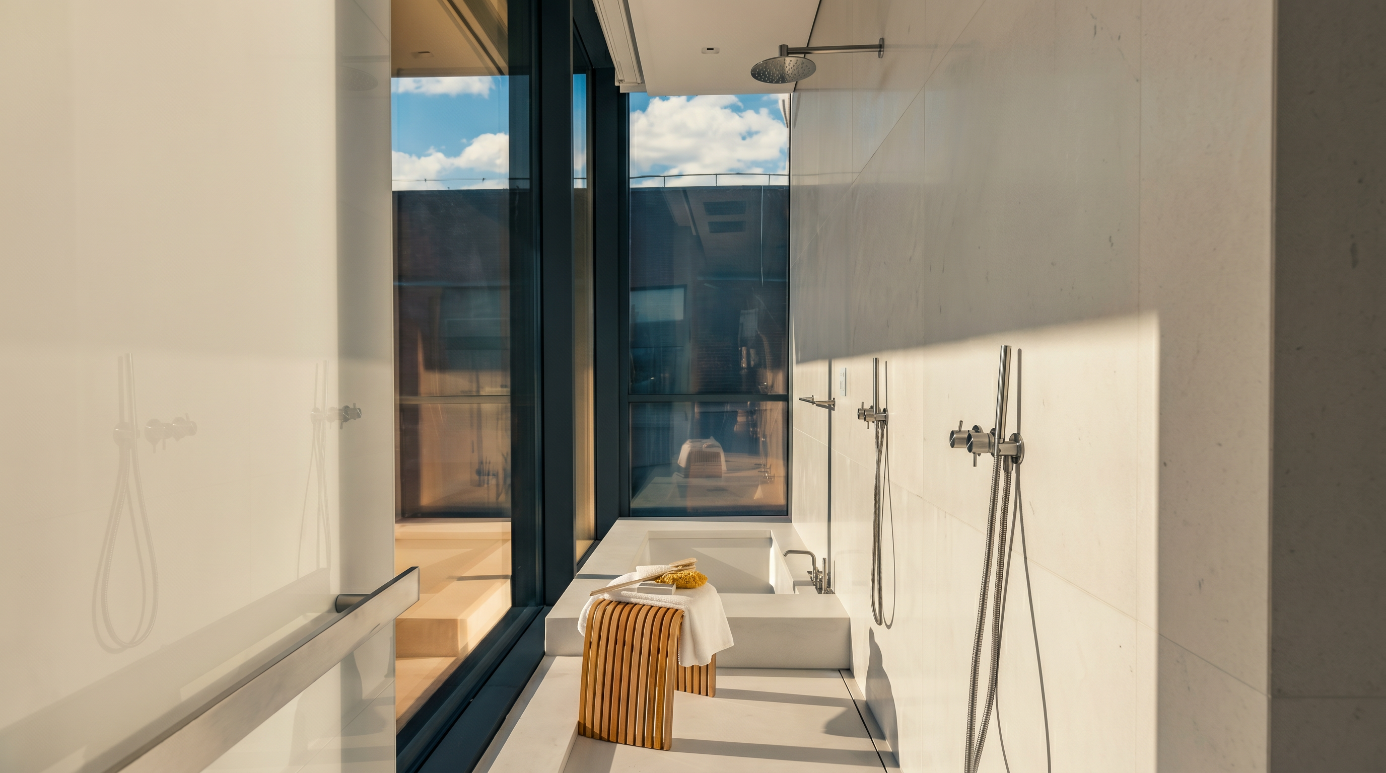 This minimalist, spa-inspired bathroom features a long, narrow layout with a built-in soaking tub at the far end and a walk-in shower area. The space is defined by clean lines, light-toned surfaces, and floor-to-ceiling windows that provide an expansive view of the outdoors. A small, slatted wood stool sits in the center, adding a warm, organic texture to the otherwise sleek and modern environment.