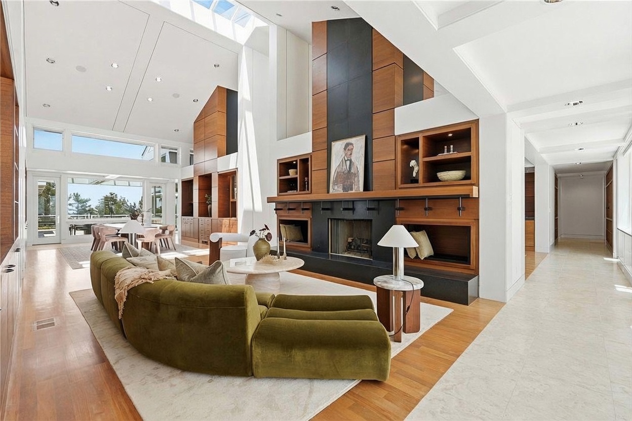 This expansive living room features a high, vaulted ceiling with large windows that flood the space with natural light. A prominent, tall fireplace with a dark surround serves as the focal point, flanked by custom wood shelving units. The room is anchored by a large, curved olive-green sofa and a circular stone coffee table, creating a sophisticated and airy atmosphere.