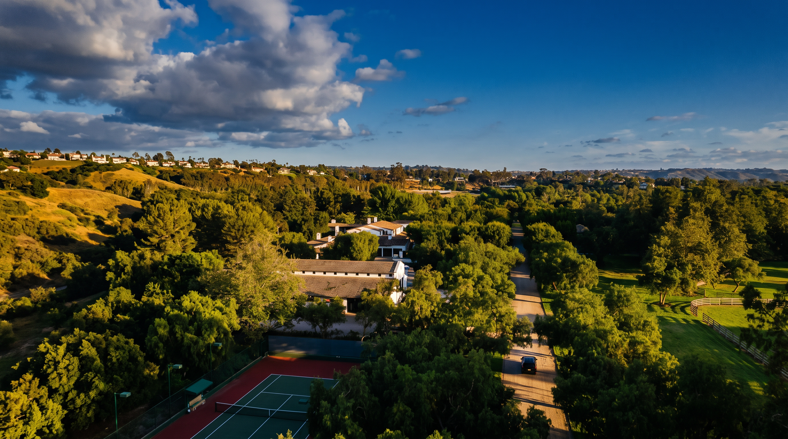 This high-angle aerial view captures a sprawling estate nestled within a lush, tree-filled landscape. The property features a long, winding driveway leading toward a traditional-style residence with a tiled roof, adjacent to a private tennis court. The expansive grounds are characterized by mature trees, open green spaces, and a peaceful, rural atmosphere.