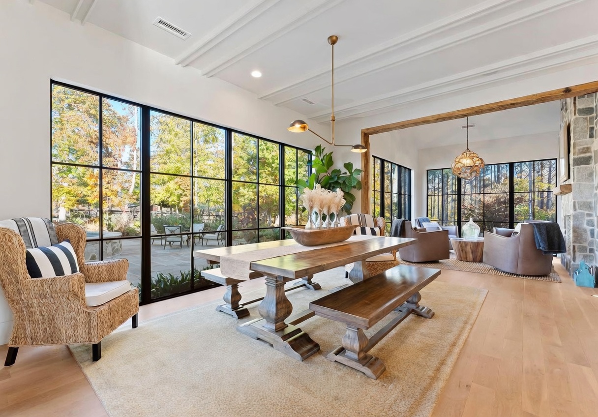 This elegant dining area features a large wooden table with a bench and wicker-style chairs, set atop a neutral area rug. The space is defined by expansive, grid-paned windows that provide abundant natural light and views of the outdoor patio, while a modern gold-toned chandelier hangs above the table. A rustic wooden beam separates the dining space from an adjacent living area, which includes comfortable seating and a stone-clad chimney breast.