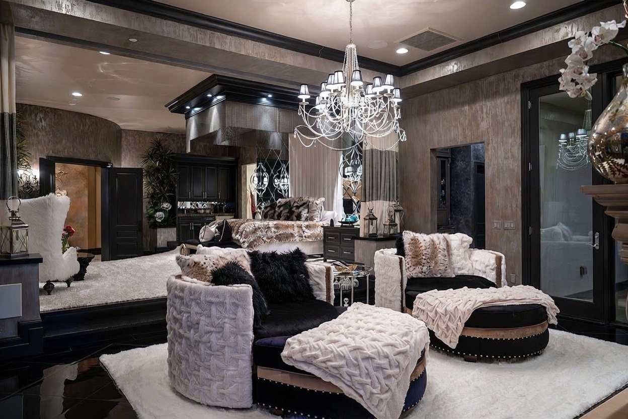 This opulent primary bedroom suite features a raised platform area housing a large bed with patterned bedding, set against a backdrop of dark cabinetry and mirrored accents. The foreground includes two plush, rounded armchairs with matching ottomans, all arranged on a soft white rug over a dark, reflective floor. A grand crystal chandelier serves as the focal point, suspended from a coffered ceiling, creating a sophisticated and dramatic atmosphere.