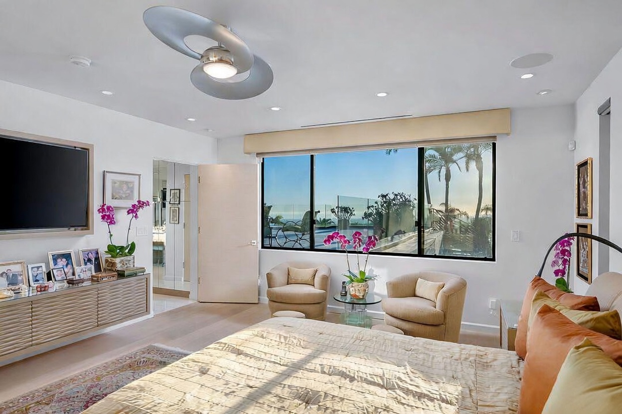 This primary bedroom features a neutral color palette with light wood flooring and a large bed dressed in textured bedding. A media console sits beneath a wall-mounted television, while two armchairs are positioned near large windows that offer a view of the outdoor deck and horizon. The room is accented by a unique, sculptural ceiling light fixture and fresh orchids, creating a serene and sophisticated atmosphere.