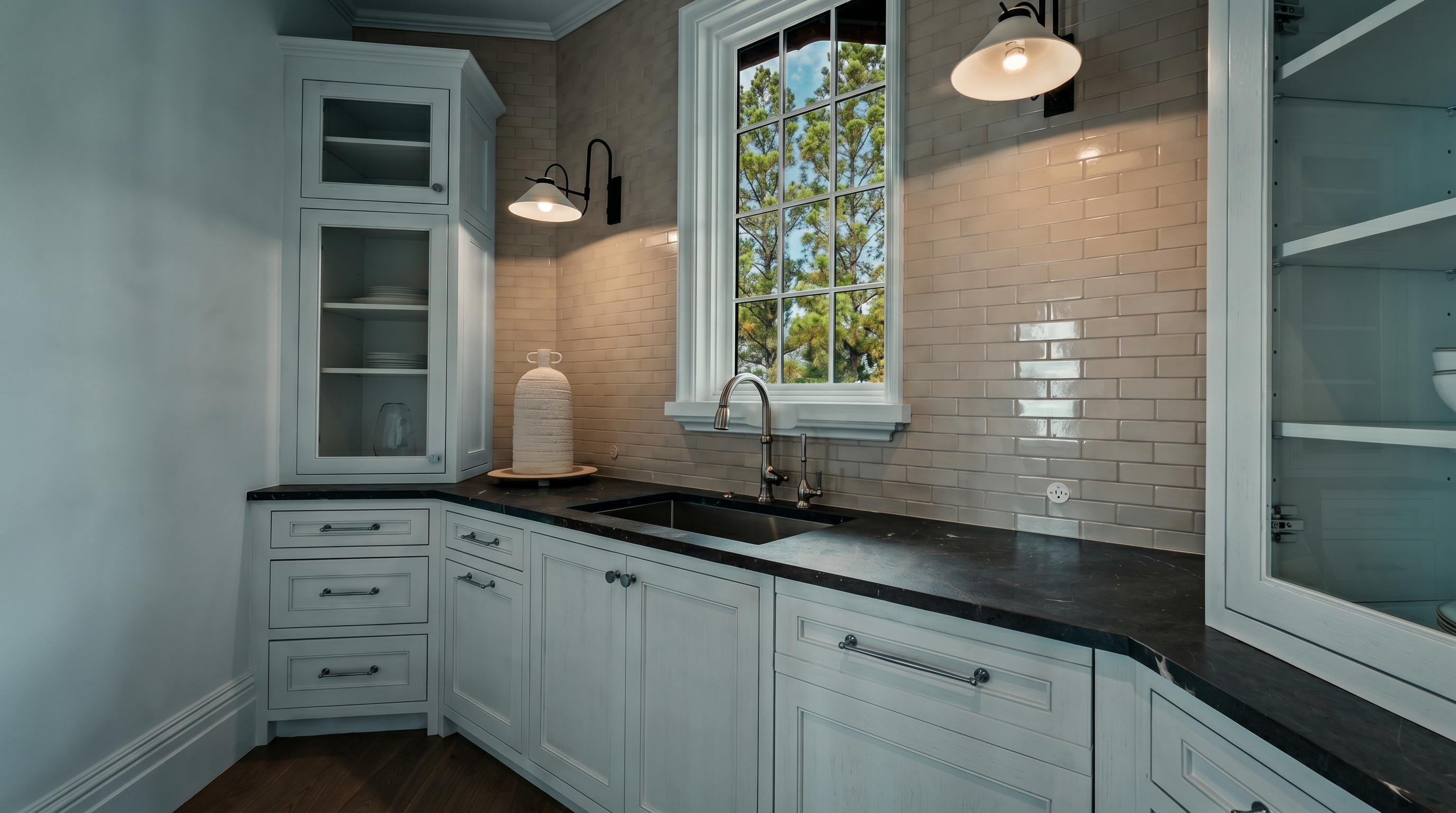 This elegant butler's pantry or secondary kitchen area features crisp white cabinetry, dark stone countertops, and a classic subway tile backsplash. A central window provides natural light, complemented by two wall-mounted sconces, while the space is finished with light-toned wood flooring. The perspective is a straight-on shot capturing the functional layout of the sink area and surrounding storage.
