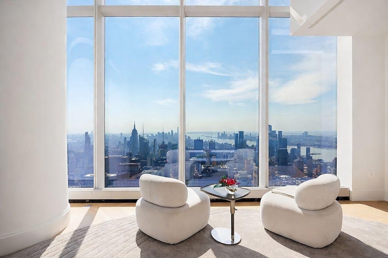 This sophisticated living space features two plush, cream-colored armchairs positioned before expansive floor-to-ceiling windows that offer a breathtaking panoramic view of the New York City skyline. A small, sleek side table with a floral arrangement sits between the chairs, resting on a neutral-toned area rug. The minimalist design emphasizes the dramatic urban backdrop, creating an atmosphere of luxury and tranquility high above the city.