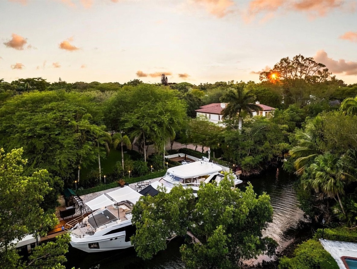 This high-angle aerial perspective captures a luxurious yacht docked along a private canal, surrounded by lush, mature tropical landscaping. The scene is bathed in the warm, golden light of a sunset, highlighting the serene waterfront setting and the elegant architecture of the nearby residence. The composition emphasizes the property's exclusive access to the water and its tranquil, secluded atmosphere.