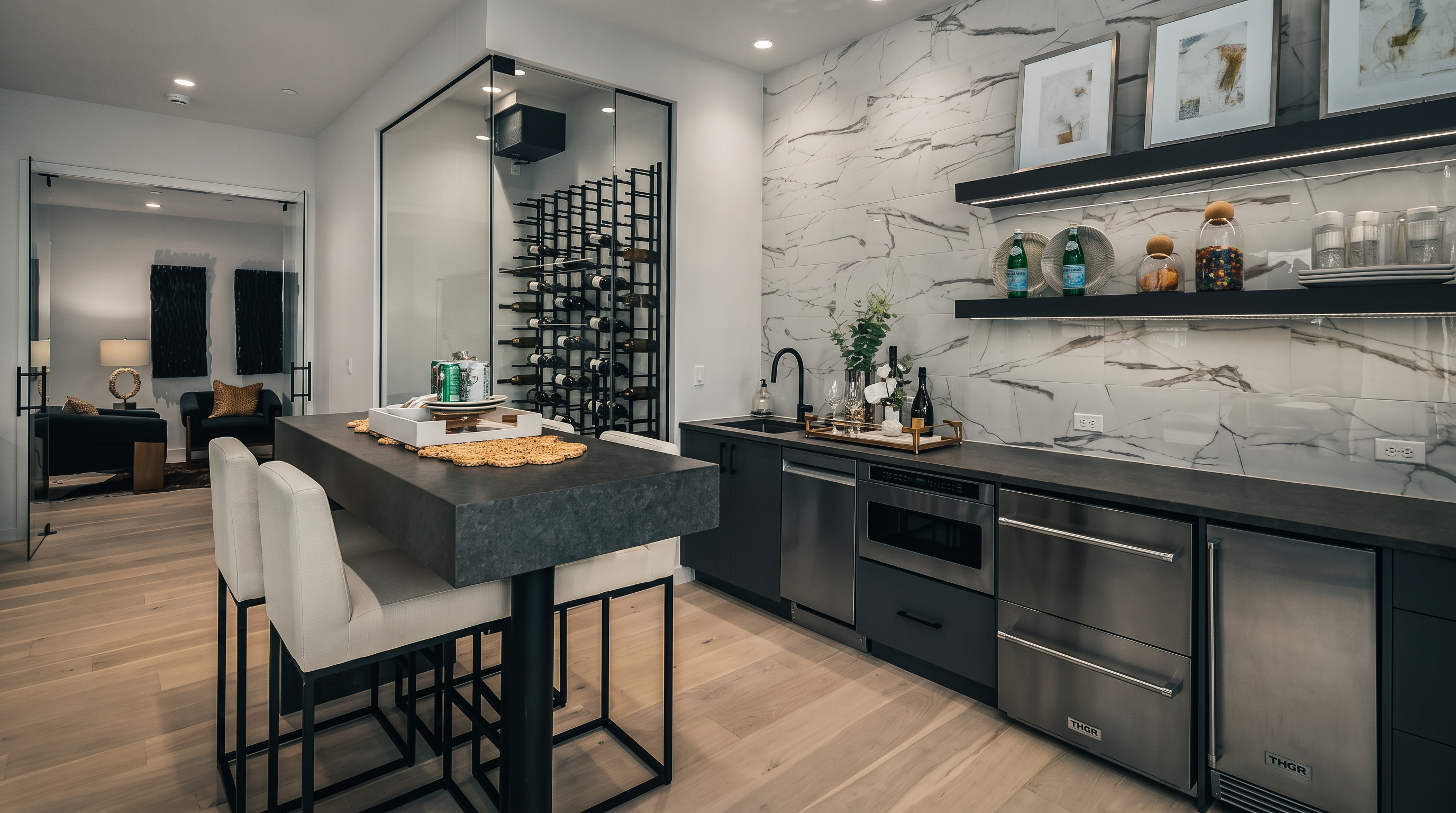 This sophisticated home bar and wine cellar area features a sleek, glass-enclosed wine storage unit with metal racking. The adjacent wet bar is equipped with stainless steel appliances, dark cabinetry, and a striking stone-patterned backsplash, complemented by two floating shelves. A dark-toned island with bar seating provides a functional focal point, all set against light-toned wood flooring in a contemporary, high-end space.