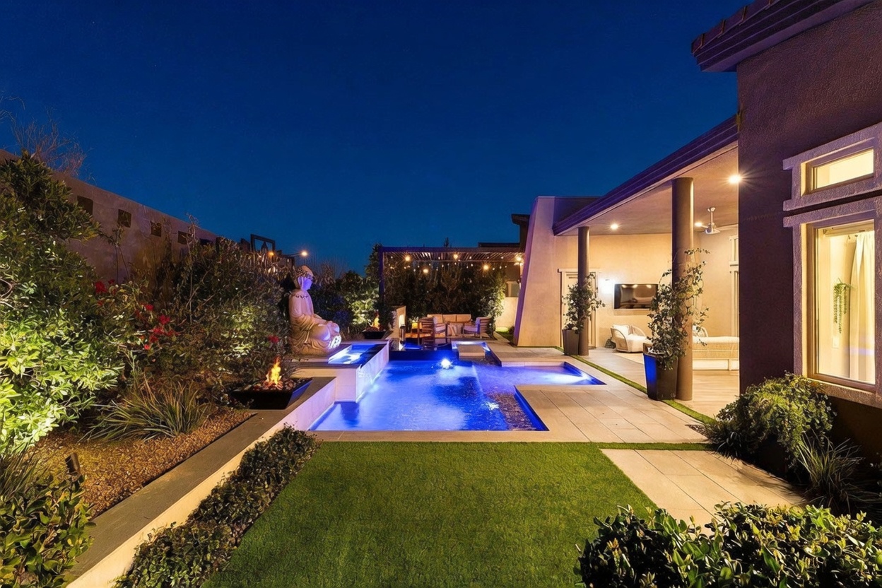 This evening shot captures a luxurious backyard oasis featuring a modern, illuminated swimming pool with integrated water features and a prominent Buddha statue. A covered patio area with comfortable seating and string lighting extends from the house, creating a seamless transition between indoor and outdoor living spaces. The scene is framed by manicured landscaping and artificial turf, offering a sophisticated and tranquil atmosphere perfect for evening entertaining.