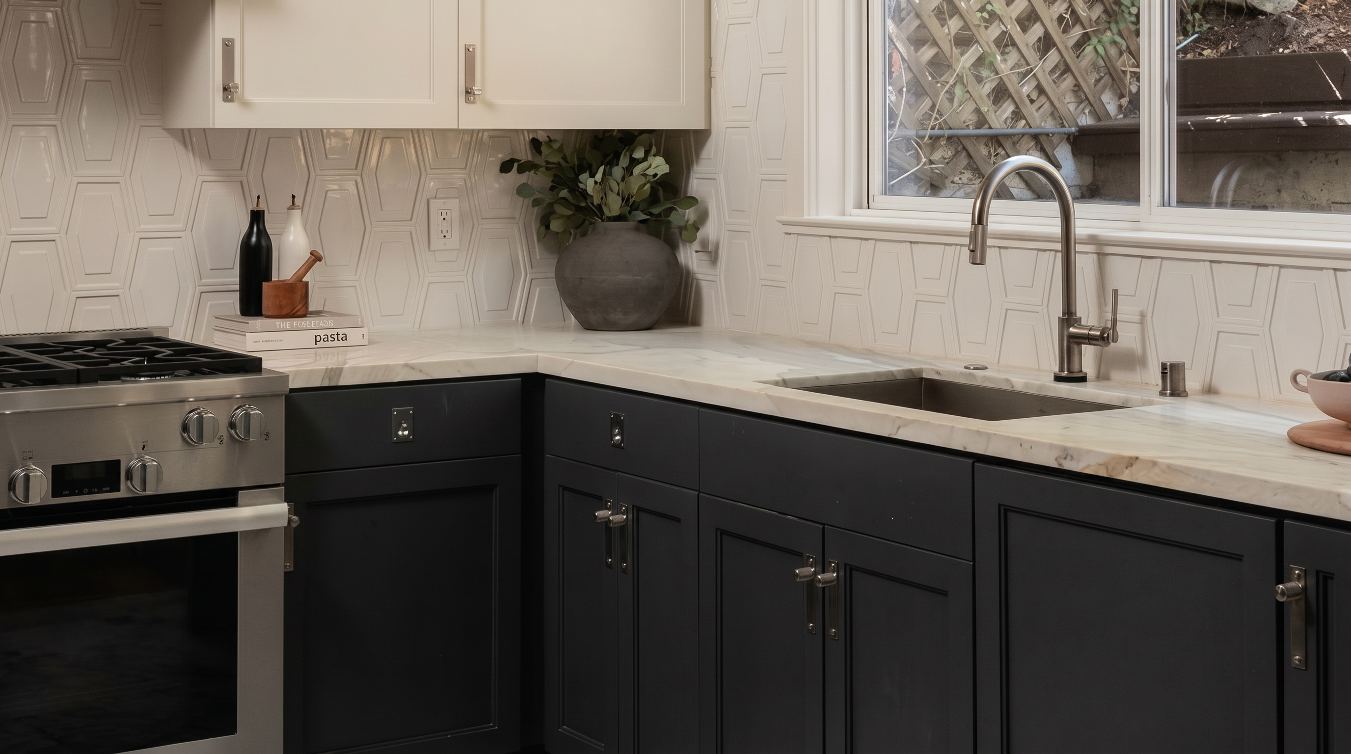 This sleek and modern kitchen features a striking contrast between dark charcoal lower cabinetry and crisp white upper cabinets. The space is anchored by a high-end stainless steel gas range, marble-look countertops, and a sophisticated white geometric tile backsplash. A deep undermount sink with a gooseneck faucet is positioned beneath a window, providing natural light and a clean, functional aesthetic.