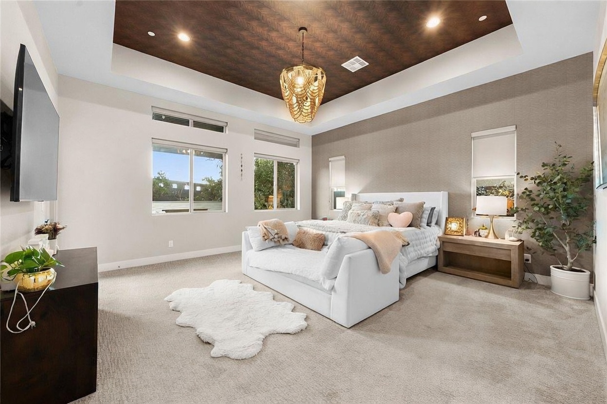 This spacious primary bedroom features a tray ceiling with a dark, textured wood-look finish and a decorative gold-toned chandelier. The room is anchored by a large white upholstered bed with a plush bench at the foot, complemented by a soft, light-colored carpet and a faux-fur rug. Large windows allow natural light to fill the space, while a tall potted plant and a dark wood dresser add contrast to the neutral color palette.