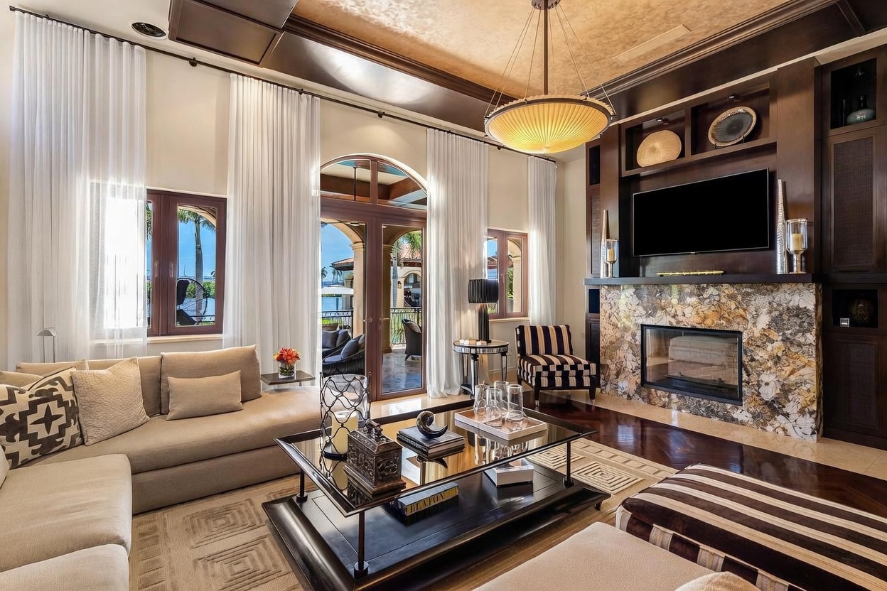 This elegant living room features a sophisticated blend of modern and traditional design, anchored by a striking floor-to-ceiling stone fireplace and a large flat-screen television. The space is bathed in natural light from floor-to-ceiling windows and glass doors that open to a scenic waterfront patio. Rich dark wood accents, a coffered ceiling, and a luxurious chandelier create a warm, high-end atmosphere perfect for entertaining.