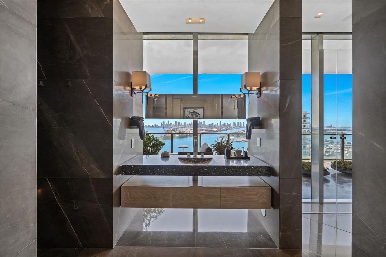 This luxurious bathroom features a floating vanity with a dark, speckled stone countertop and a light wood base, centered against a wall of large, dark, polished stone tiles. A mirror reflects the expansive, panoramic view of the city skyline and harbor visible through the floor-to-ceiling windows behind the vanity. The space is framed by symmetrical wall sconces and polished stone flooring, creating a sophisticated and high-end atmosphere.