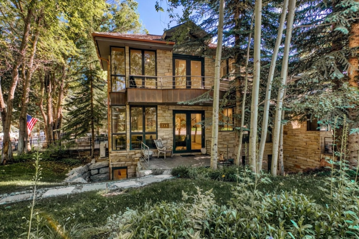 This multi-level home features a striking exterior clad in textured stone and accented with dark-framed windows and copper-toned metal detailing. The structure includes a second-story balcony with a sleek metal railing and a ground-level patio area nestled among mature aspen trees. The natural, wooded setting provides a serene backdrop, emphasizing the home's integration with its forest environment.