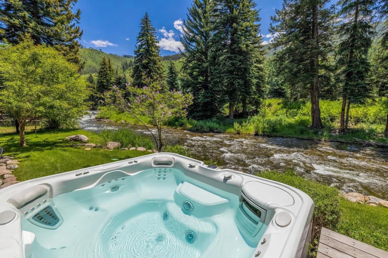 This image captures a relaxing outdoor spa experience, featuring a modern, light-colored hot tub positioned on a deck overlooking a rushing mountain stream. The lush, green landscape is filled with tall pine trees and vibrant foliage, creating a serene and private natural setting. The perspective is from just behind the spa, drawing the viewer's eye directly toward the scenic river and the forested hillside beyond.