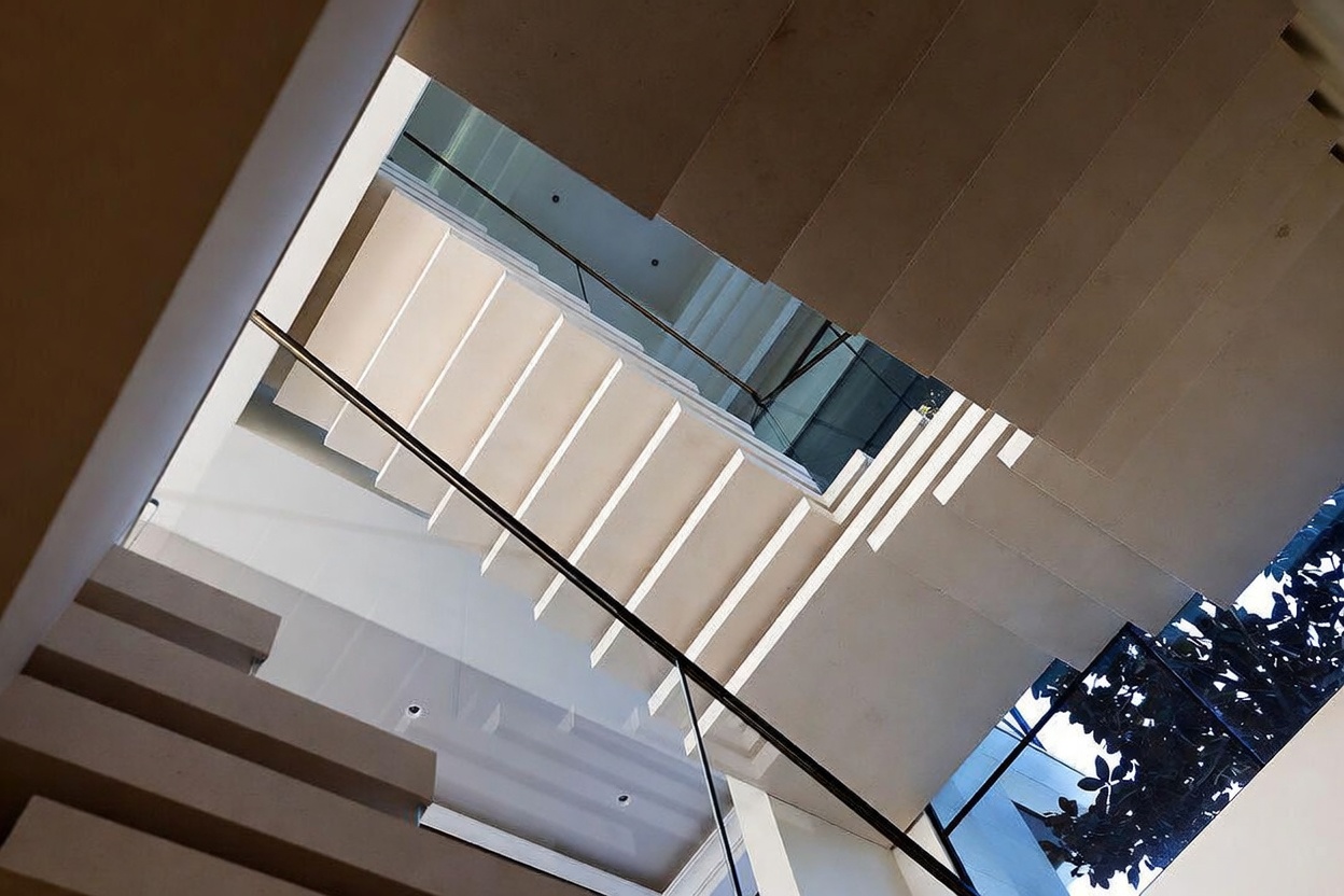 This low-angle, upward-looking perspective captures a modern staircase featuring floating stone treads and a sleek glass railing. The design emphasizes clean lines and geometric shapes, with natural light filtering through the overhead structure to create a bright, airy atmosphere. The minimalist aesthetic highlights the architectural precision of the staircase as it ascends through the open space.