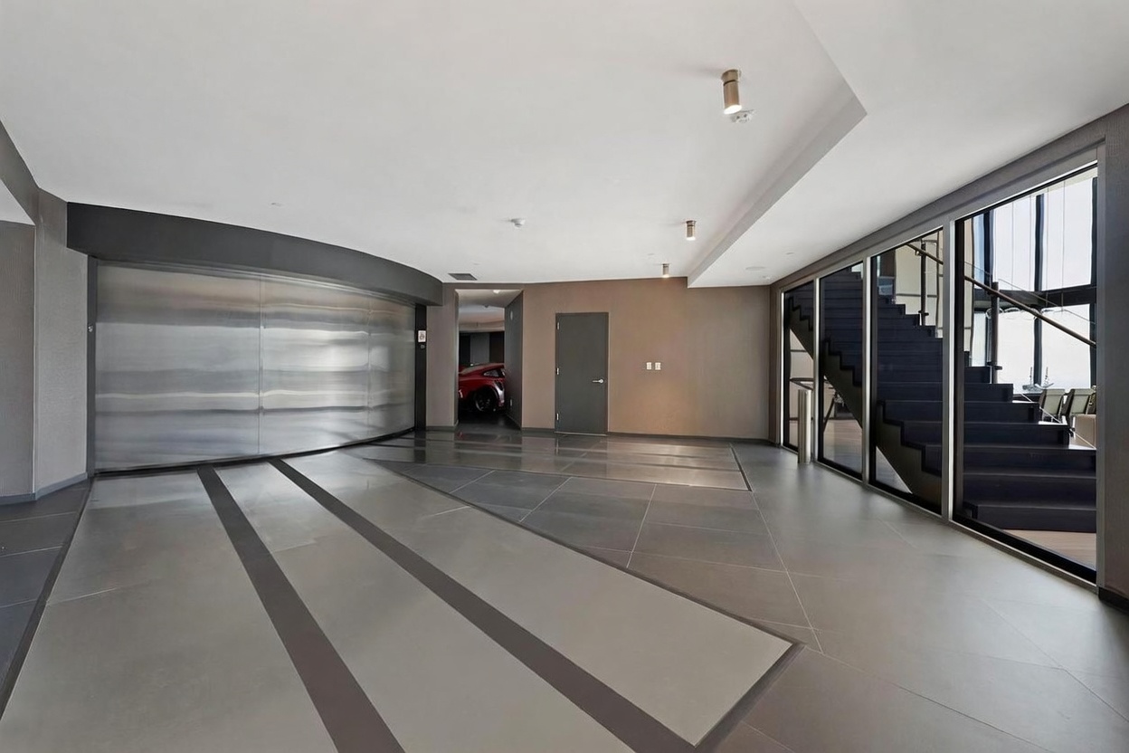 This image showcases a high-end, modern private garage featuring a large, curved metallic door and sleek, dark-toned tile flooring with integrated lighting strips. A staircase with glass railings is visible to the right, leading to an upper level, while a glimpse of a red sports car can be seen through an open doorway in the background. The space is minimalist and industrial in style, emphasizing a sophisticated, gallery-like aesthetic for vehicle storage.