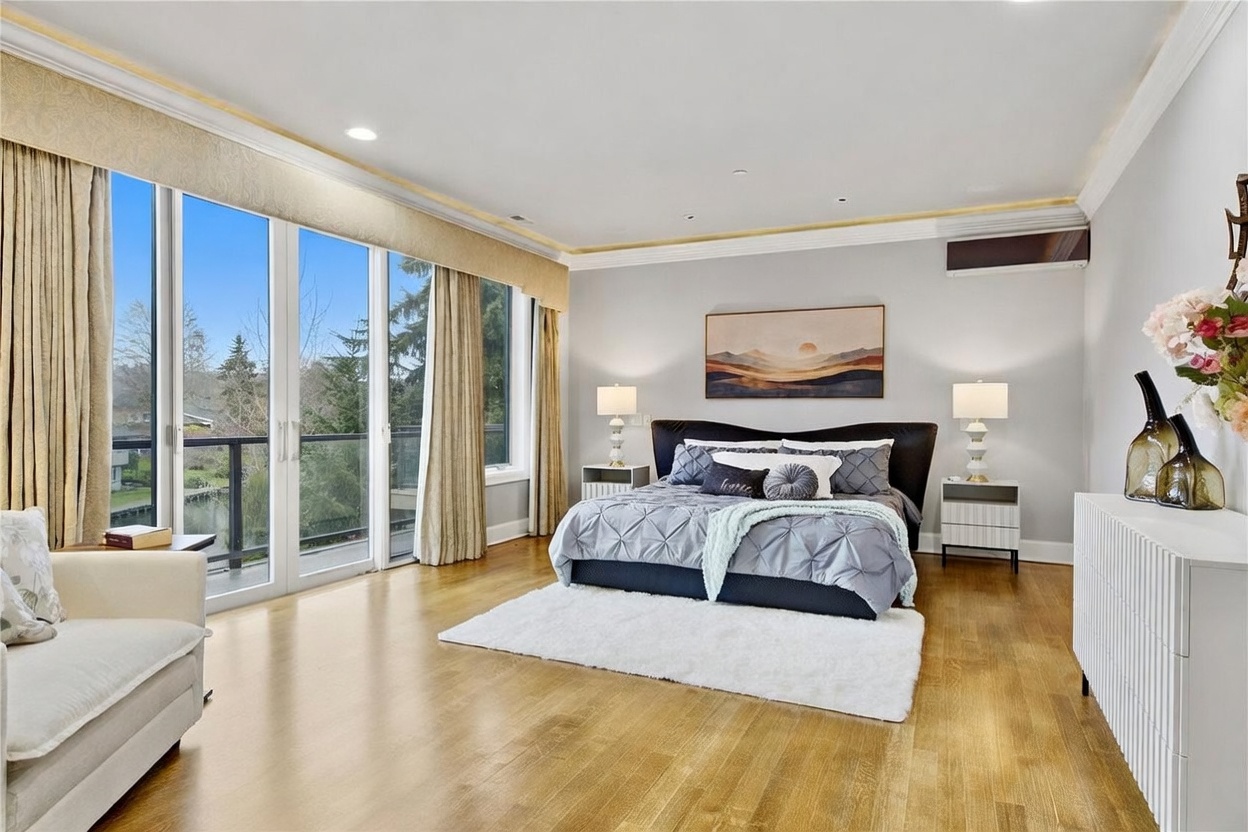 This spacious primary bedroom features light wood flooring and a neutral color palette, centered around a large bed with a dark upholstered headboard. A wall of floor-to-ceiling windows and sliding glass doors provides abundant natural light and access to a private balcony overlooking the outdoors. The room is accented by a landscape painting above the bed, matching bedside tables with lamps, and a white dresser topped with decorative vases.