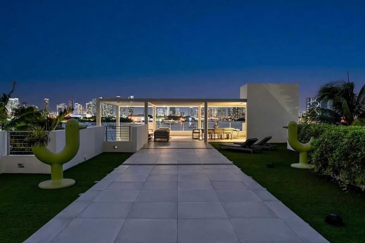 This expansive rooftop terrace offers a sophisticated outdoor living space with a central tiled walkway flanked by artificial turf and whimsical green sculptural accents. The area features a covered dining and lounge section, providing a seamless transition to panoramic views of the city skyline and waterfront at night. The modern design emphasizes clean lines and an open-air atmosphere, perfect for evening entertaining.
