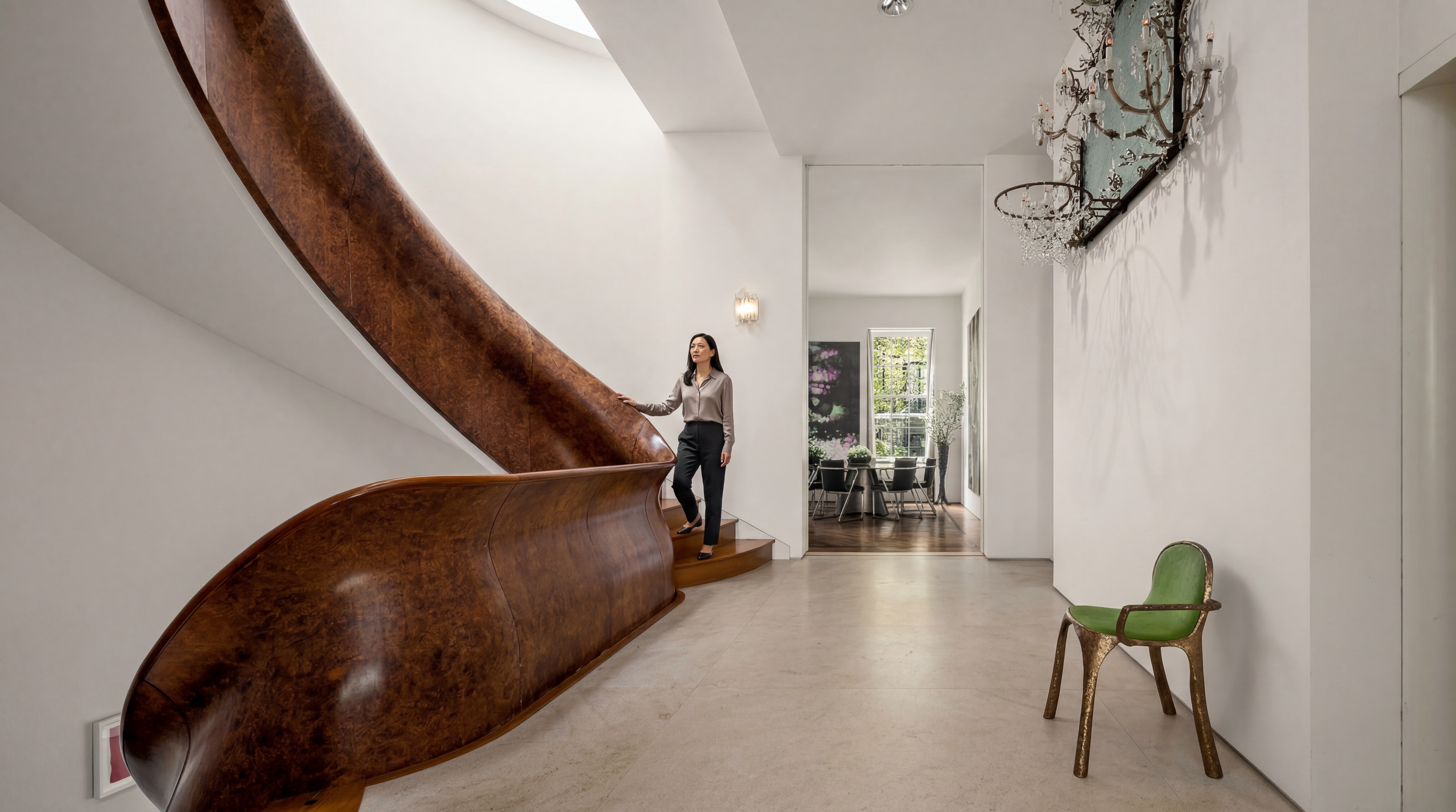 This elegant entryway features a striking, curved staircase with a prominent, polished wood-grain banister that sweeps down into the foyer. The space is characterized by minimalist white walls, light-toned stone flooring, and a unique green accent chair positioned near a decorative crystal chandelier. A clear line of sight leads through an open doorway into a bright dining area, creating a sense of depth and architectural sophistication.